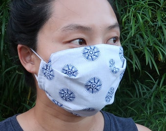 Puede incluir: Una persona lleva una mascarilla facial blanca con un patrón de brújula azul. La mascarilla está plisada y tiene lazos elásticos para las orejas. El texto "Blue Nautical" y "DIAMOND CITY CO." es visible en la parte inferior de la imagen.