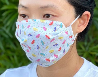 Puede incluir: Una mascarilla facial blanca con un patrón de donuts coloridos. La mascarilla tiene una banda elástica blanca y el texto "Donuts" está impreso debajo de la mascarilla.