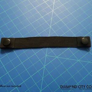 K&ouml;nnte beinhalten: Schwarzes Gummiband mit zwei schwarzen Kn&ouml;pfen an jedem Ende. Das Band liegt auf einem blauen Gitterhintergrund. Maske nicht im Lieferumfang enthalten. DIAMOND CITY CO.