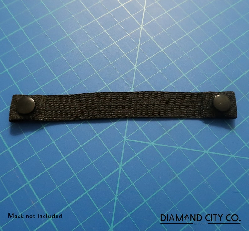 K&ouml;nnte beinhalten: Schwarzes Gummiband mit zwei schwarzen Kn&ouml;pfen an jedem Ende. Das Band liegt auf einem blauen Gitterhintergrund. Maske nicht im Lieferumfang enthalten. DIAMOND CITY CO.