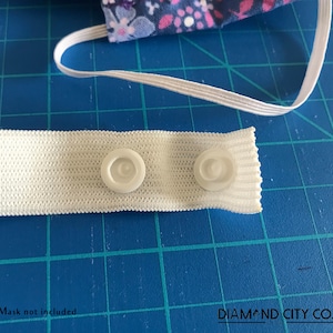 K&ouml;nnte beinhalten: Wei&szlig;es Gummiband mit zwei wei&szlig;en Plastikkn&ouml;pfen. Das Band liegt auf einer blauen Schneidematte. Maske nicht im Lieferumfang enthalten. DIAMOND CITY CO.