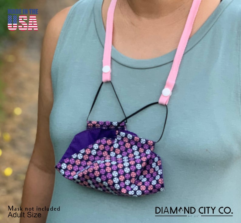 Puede incluir: Cord&oacute;n rosa para mascarilla facial con clips de pl&aacute;stico blancos. El cord&oacute;n est&aacute; hecho en los Estados Unidos. El texto "DIAMOND CITY CO. EST 2017" es visible en la esquina inferior derecha de la imagen.