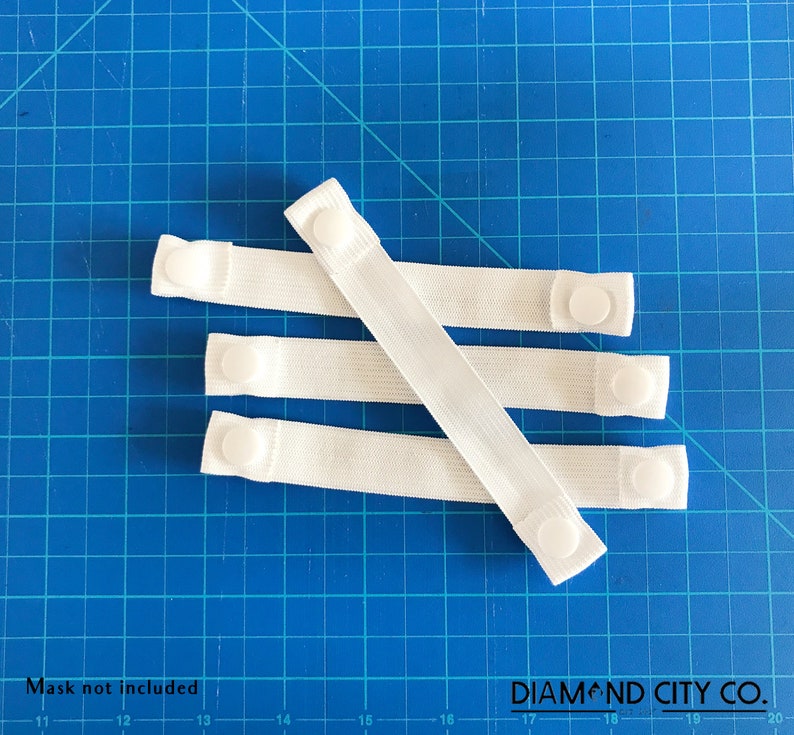 K&ouml;nnte beinhalten: Vier wei&szlig;e elastische B&auml;nder mit wei&szlig;en Plastikkn&ouml;pfen an jedem Ende. Die B&auml;nder liegen auf einer blauen, karierten Oberfl&auml;che. Der Text "DIAMOND CITY CO." ist auf der Oberfl&auml;che aufgedruckt.