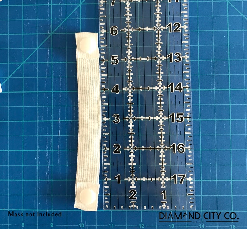 K&ouml;nnte beinhalten: Wei&szlig;es Gummiband mit zwei wei&szlig;en Plastikkn&ouml;pfen. Das Band wird an einem Lineal gemessen und ist etwa 7,6 cm lang.