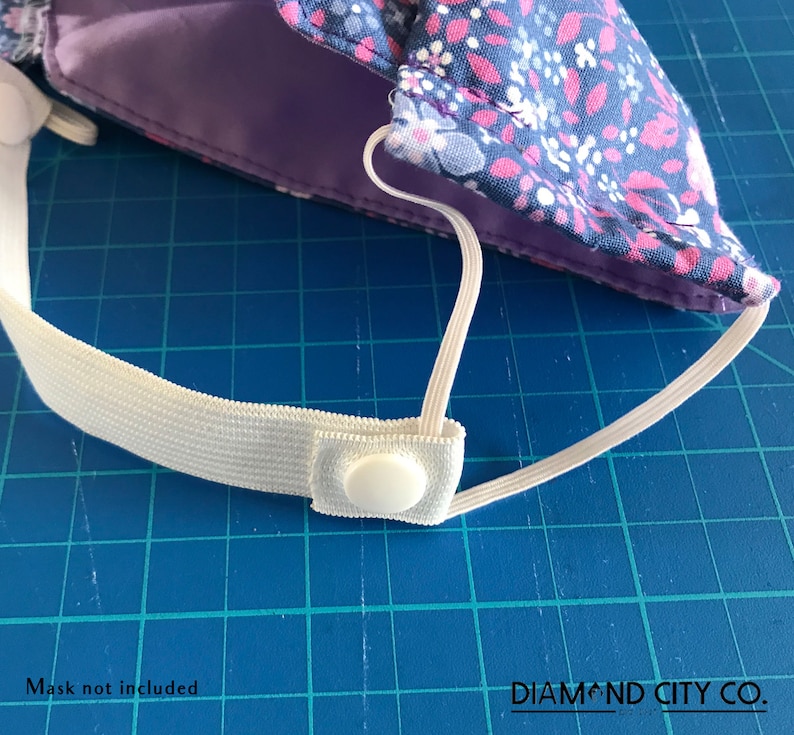 K&ouml;nnte beinhalten: Wei&szlig;es Gummiband mit einem wei&szlig;en Plastikknopf. Das Band ist an einer blauen und pinken floralen Stoffmaske befestigt. Der Text "DIAMOND CITY CO." ist in der rechten unteren Ecke des Bildes sichtbar.