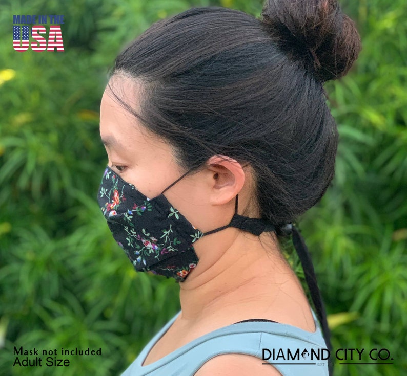 Puede incluir: Una mascarilla facial negra con estampado floral y correas ajustables. La mascarilla est&aacute; hecha en los Estados Unidos y es de talla para adultos. El texto "DIAMOND CITY CO." es visible en la esquina inferior derecha de la imagen.