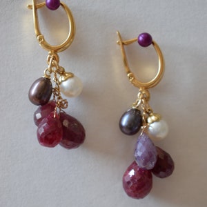 May include: Gold earrings with a purple bead at the top and dangling charms. The charms include a white pearl, a dark purple pearl, and three red ruby stones.
