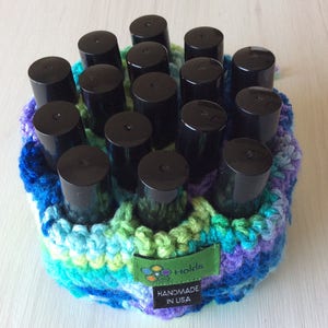 May include: A handmade, crocheted nail polish holder in shades of blue, green, and purple. The holder contains multiple black nail polish bottles. A green tag reads "Holds" and "HANDMADE IN USA."