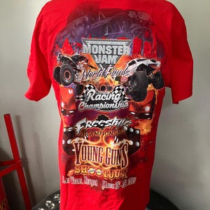 Puede incluir: Camiseta roja con un gr&aacute;fico de Monster Jam World Finals que presenta camiones monstruo, llamas y el texto "Young Guns Shootout" con la fecha "March 26-28 2015" y la ubicaci&oacute;n "Las Vegas, Nevada".