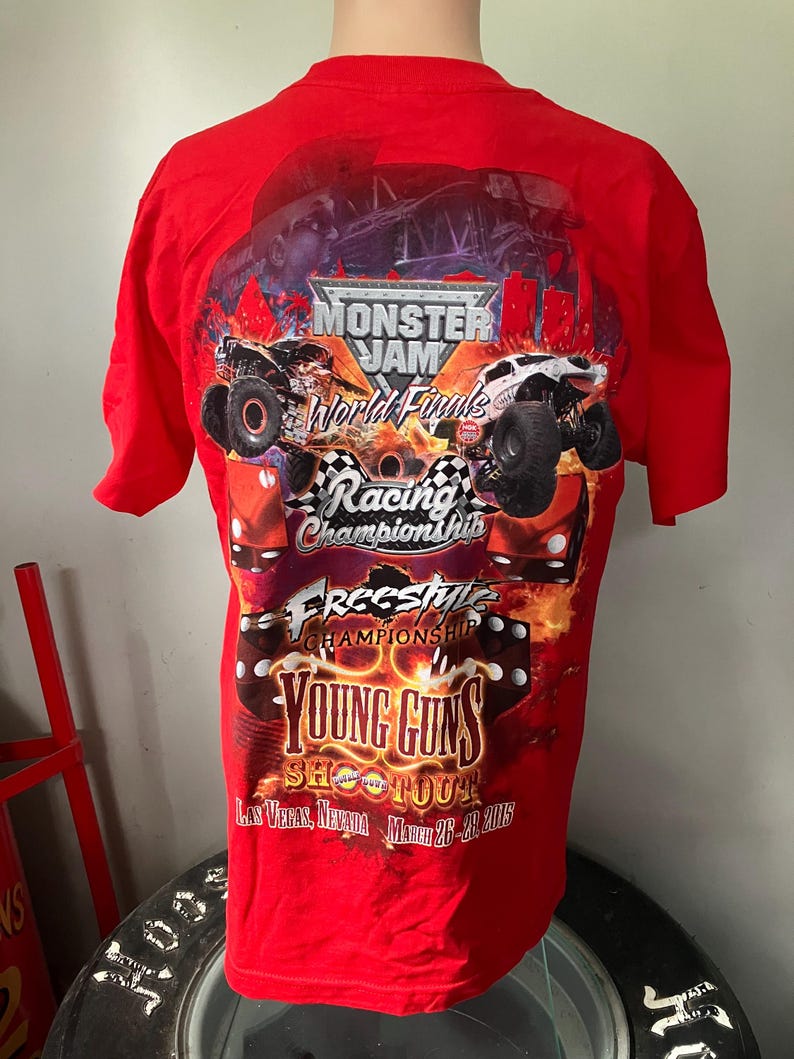 Puede incluir: Camiseta roja con un gr&aacute;fico de Monster Jam World Finals que presenta camiones monstruo, llamas y el texto "Young Guns Shootout" con la fecha "March 26-28 2015" y la ubicaci&oacute;n "Las Vegas, Nevada".