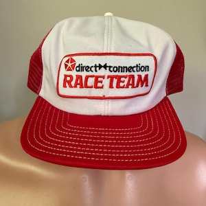Pode incluir: Um boné de caminhoneiro Direct Connection Race Team branco e vermelho. O boné apresenta um painel frontal branco com um patch vermelho que diz "direct connection RACE TEAM" em letras vermelhas. A aba é vermelha com costuras brancas e as laterais de malha são vermelhas.