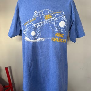 Może przedstawiać: Niebieska koszulka z biało-żółtą grafiką monster trucka. Projekt zawiera tekst "Auto Value", "Parts Master" i "King Krunch". Koszulka jest prezentowana na manekinie.