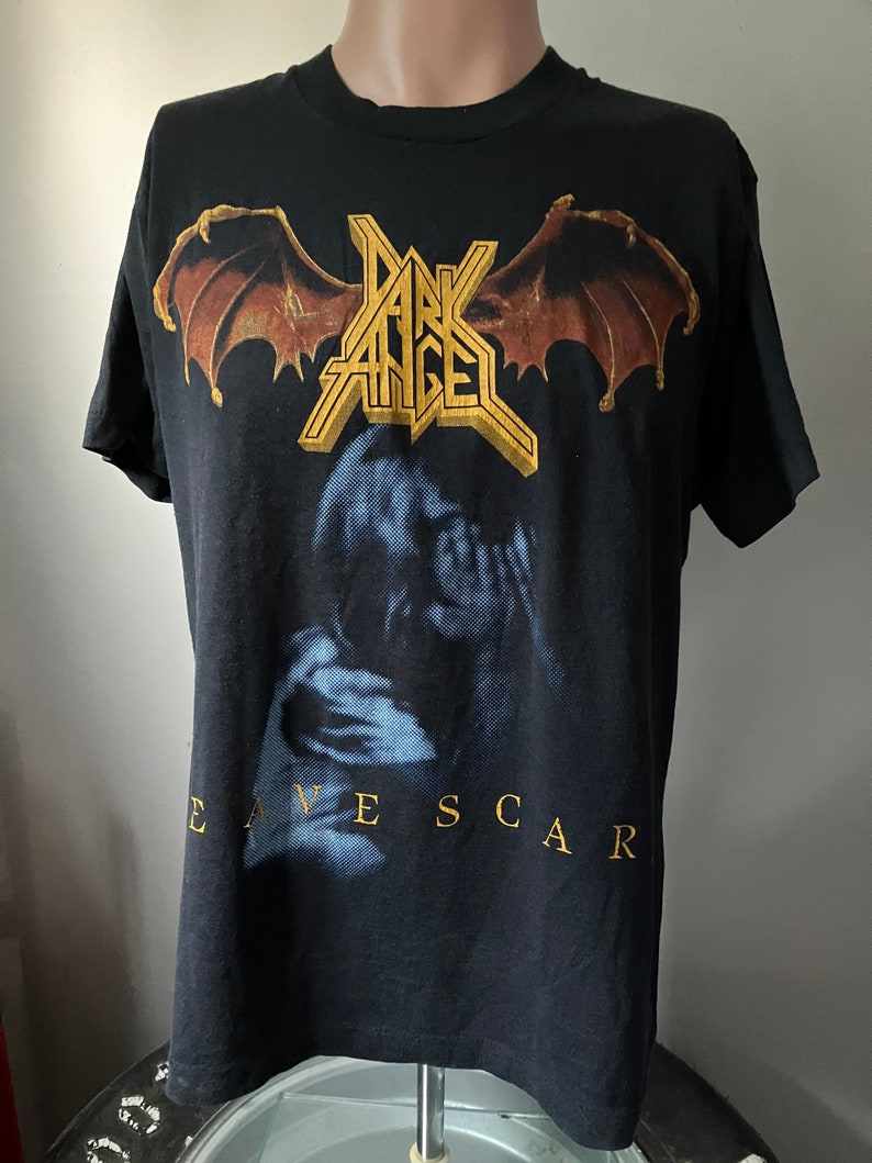 Puede incluir: Camiseta negra con un gr&aacute;fico dorado y marr&oacute;n de una banda llamada "Dark Angel" con las palabras "Eavescar" impresas debajo del gr&aacute;fico.