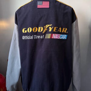 May include: A black and gray jacket with a Goodyear logo and the words "Official Tire of NASCAR" on the back. The jacket also has a small American flag patch on the upper back.
