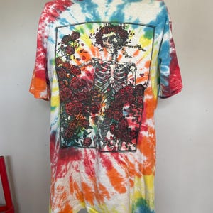 May include: Tie-dye t-shirt with a skeleton graphic surrounded by red roses. The shirt features a vibrant swirl of colors including red, orange, yellow, blue, and white. The graphic is framed in a black rectangle.