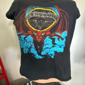 May include: Black t-shirt featuring a graphic of a red devil with wings, a pentagram, and the text "BLACK SABBATH" in a yellow and black circle. The design includes blue accents. The shirt is displayed on a mannequin.