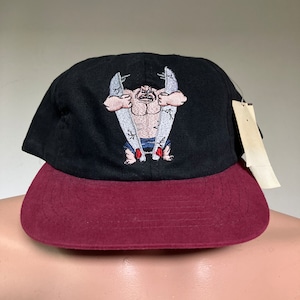 May include: Black and maroon baseball cap with an embroidered image of a cartoon character holding two surfboards.