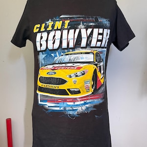 Peut inclure: Un t-shirt noir à manches courtes avec un imprimé représentant une voiture de course Ford Fusion jaune. Le texte "CLINT BOWYER" est inscrit en grandes lettres blanches et jaunes au-dessus de la voiture, avec des étoiles rouges, blanches et bleues.