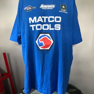 Può includere: T-shirt blu con la scritta "MATCO TOOLS" in lettere bianche. La maglietta presenta i loghi di "Aaron's Dream Machine", "U.S. Army" e "Full Throttle". Un logo rosso e bianco è centrato sotto.