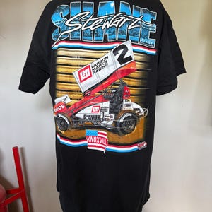 Puede incluir: Camiseta negra con un diseño de coche de carreras, con el nombre "Shane Stewart" en azul y blanco. El diseño incluye el número 2, las palabras "Larson Marks Racing" y la palabra "Knoxville".