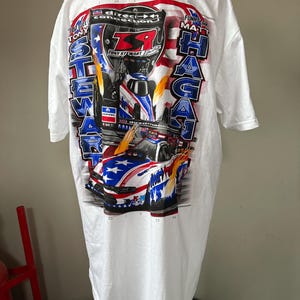 Pode incluir: Camiseta branca com um gráfico de carro de corrida, o texto "Tony Stewart" e "Matt Hagan". O design inclui a bandeira americana e o número 14. A camisa é uma edição limitada.