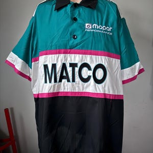 Può includere: Una camicia da lavoro a maniche corte con una sezione superiore verde acqua, una striscia bianca e rosa sul petto e una sezione inferiore nera. La camicia presenta il logo "MATCO" e il logo "mopar PERFORMANCE".
