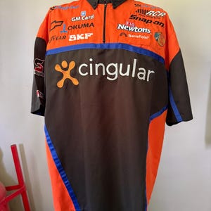May include: Orange, black, and blue racing team shirt with multiple sponsor logos, including "Cingular," "GM Card," and "Snap-on." The shirt has a black collar and sleeves, with orange and blue accents. The shirt is displayed on a mannequin.