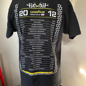 Pode incluir: Camiseta preta com um design de bandeira xadrez branca e amarela e o texto "GOODYEAR Official Tire of NHRA" com uma lista de eventos de corrida e datas.