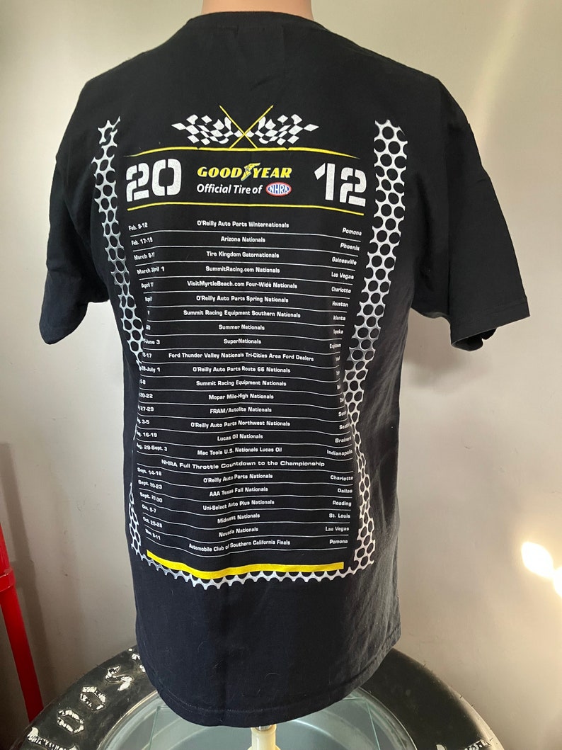 Pode incluir: Camiseta preta com um design de bandeira xadrez branca e amarela e o texto "GOODYEAR Official Tire of NHRA" com uma lista de eventos de corrida e datas.