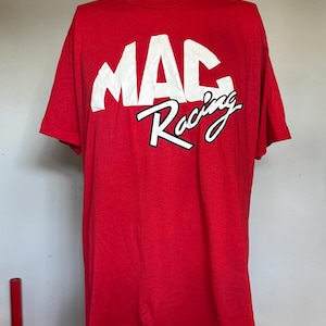 May include: A bright red short-sleeved t-shirt featuring the text MAC in large white block letters and the word Racing in a white cursive script with a black outline printed across the center of the chest on a plain red fabric background.