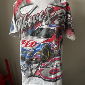 May include: White t-shirt featuring a race car design with red, blue, and gray accents. The shirt has the words "Above & Beyond" and the signature "Mark Martin" printed on it. The design includes a car with the number 44.