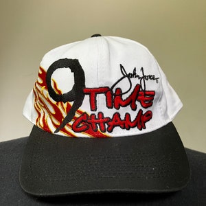 May include: White baseball cap with a black brim. The front of the cap features a red and yellow flame design with the number "9" and the text "TIME CHAMP" in black. The signature "John Force" is embroidered in black on the right side of the cap.