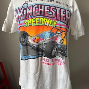May include: White t-shirt featuring a colorful graphic of a race car with the text "Winchester Speedway" and "World's Fastest Half Mile." The shirt also includes "Old Timers Weekend 1991" text. The design has a retro aesthetic.