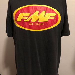 Op de afbeelding: Zwart T-shirt met een geel en rood ovaal logo met de tekst "FMF SO. CALIF."
