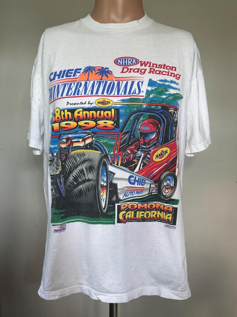 May include: A white short-sleeve t-shirt featuring a graphic for the 1998 38th Annual Chief Auto Parts NHRA Winston Drag Racing Internationals in Pomona, California, depicting a red dragster, palm trees, and the Pennzoil logo.