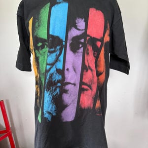 May include: Black t-shirt featuring a colorful graphic of faces in vertical stripes. The faces are segmented into vibrant colors: green, blue, purple, and red. The shirt has short sleeves and a crew neck.