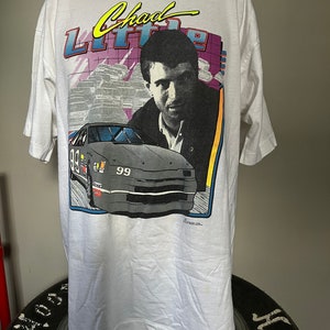 May include: White t-shirt featuring a graphic of a race car and a portrait of Chad Little. The design includes the text "Chad Little" in yellow and blue, with the number "99" on the car. The shirt is displayed on a mannequin.
