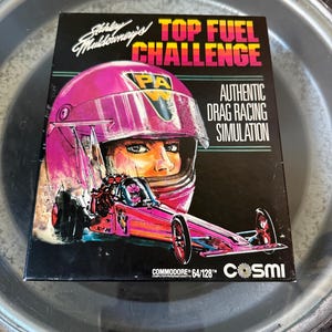 May include: A vintage video game box for "Shirley Muldowney's Top Fuel Challenge." The box art features a pink helmet and dragster, with the text "Authentic Drag Racing Simulation." The game is for Commodore 64/128.