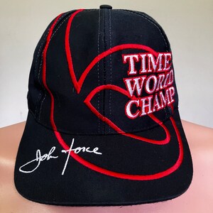 May include: Black baseball cap with red stitching and a white embroidered logo that says "Time World Champ". The cap has a white signature that says "John Force" on the front.