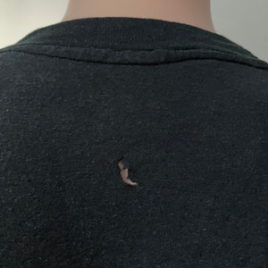 Puede incluir: Camiseta negra con un peque&ntilde;o agujero en la tela. La camiseta tiene un patr&oacute;n descolorido y moteado.