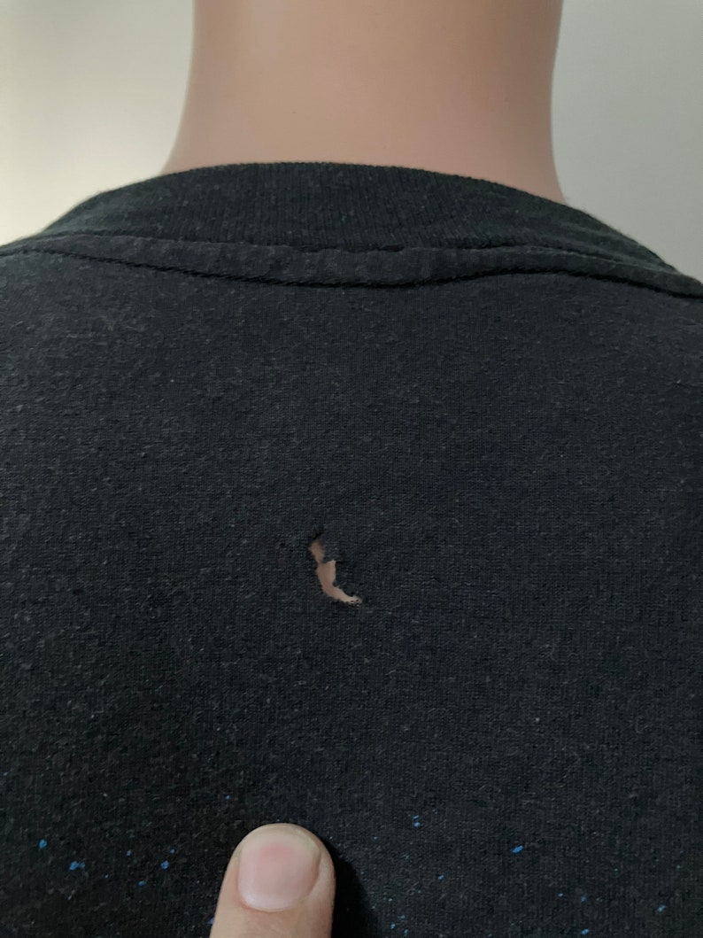 Puede incluir: Camiseta negra con un peque&ntilde;o agujero en la tela. La camiseta tiene un patr&oacute;n descolorido y moteado.