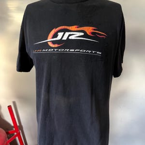May include: Black t-shirt with the JR Motorsports logo in orange and white. The logo features stylized text and a flame design. The words "JR MOTORSPORTS" are printed below the logo in white.