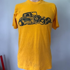 Op de afbeelding: Mosterdgeel t-shirt met een zwarte afbeelding van een vintage hot rod auto. Het autodesign bevat de tekst "Lou Settelmaier". Het shirt heeft korte mouwen en een ronde hals. Het t-shirt wordt op een mannequin getoond.