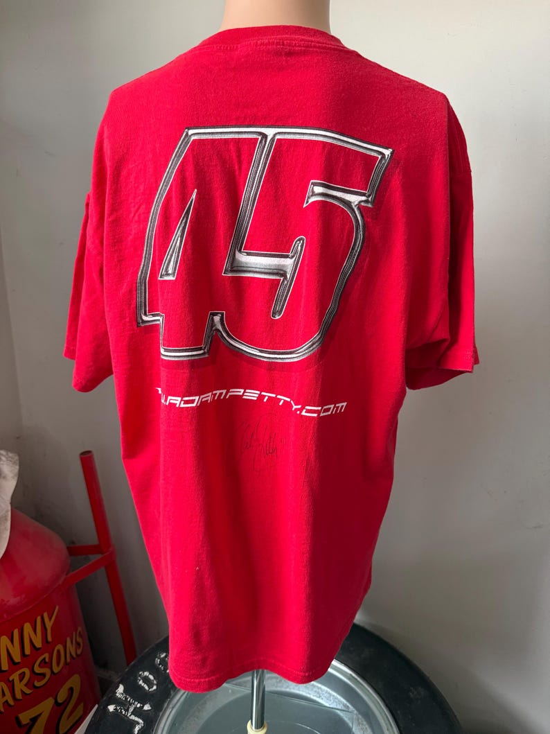 May include: Red t-shirt featuring a large silver and white number 45 on the back. The text "adam petty.com" is printed below the number. The shirt is displayed on a mannequin.