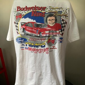 May include: White t-shirt with a graphic of a red Ford Tempo race car with the text "Budweiser King" and "260.11 mph". The graphic also includes the driver's name, "Tommy Kendall", and the sponsors "Ford", "Tempo", "Motorcraft", and "Kangaroos".