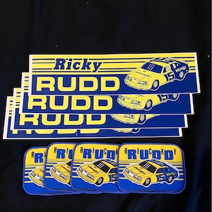 May include: Four blue and yellow stickers with the text "Ricky Rudd" and a yellow race car with the number 15 and the word "Wrangler" on it. Five blue and yellow patches with the same image and text.