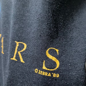 Puede incluir: Primer plano de una camiseta negra con letras doradas que dicen "R.S. &copy;ZEBRA '89".