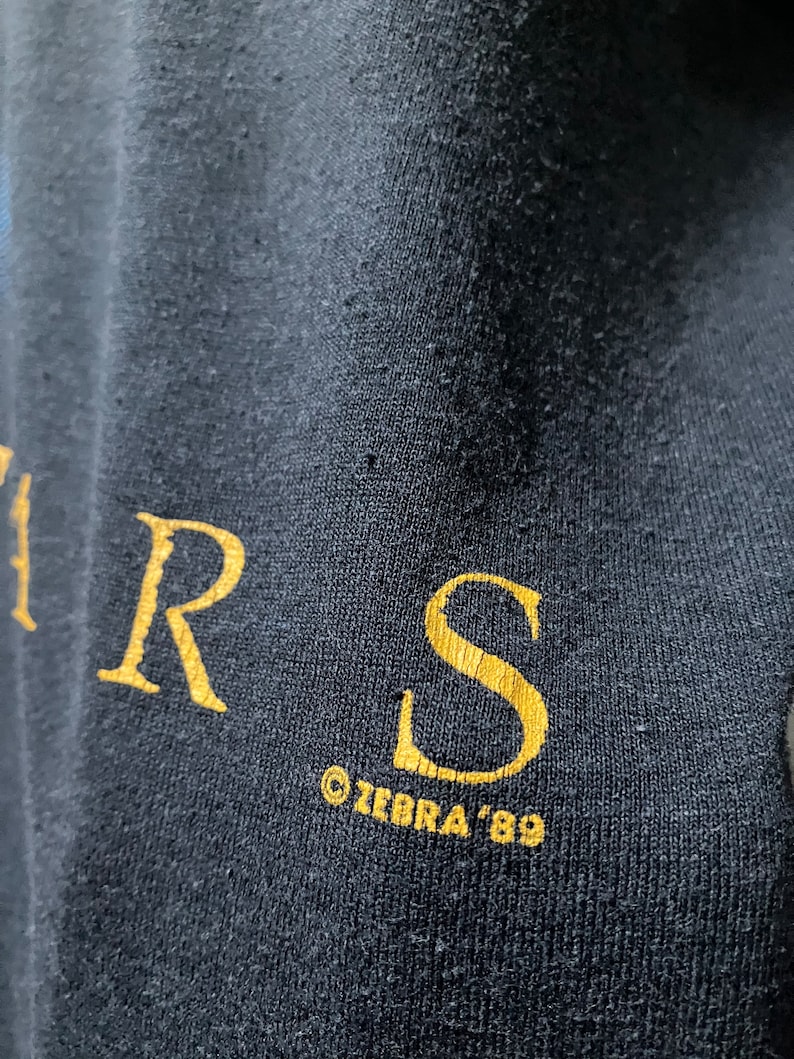 Puede incluir: Primer plano de una camiseta negra con letras doradas que dicen "R.S. &copy;ZEBRA '89".