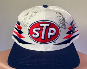 Könnte beinhalten: Weiße Baseballkappe mit einem roten und blauen STP-Logo. Die Kappe hat einen dunkelblauen Schirm und ist von mehreren Personen signiert.