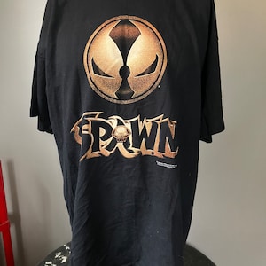 Puede incluir: Camiseta negra con un gráfico dorado del personaje Spawn. El gráfico presenta un círculo con una calavera en su interior y la palabra "Spawn" escrita debajo.